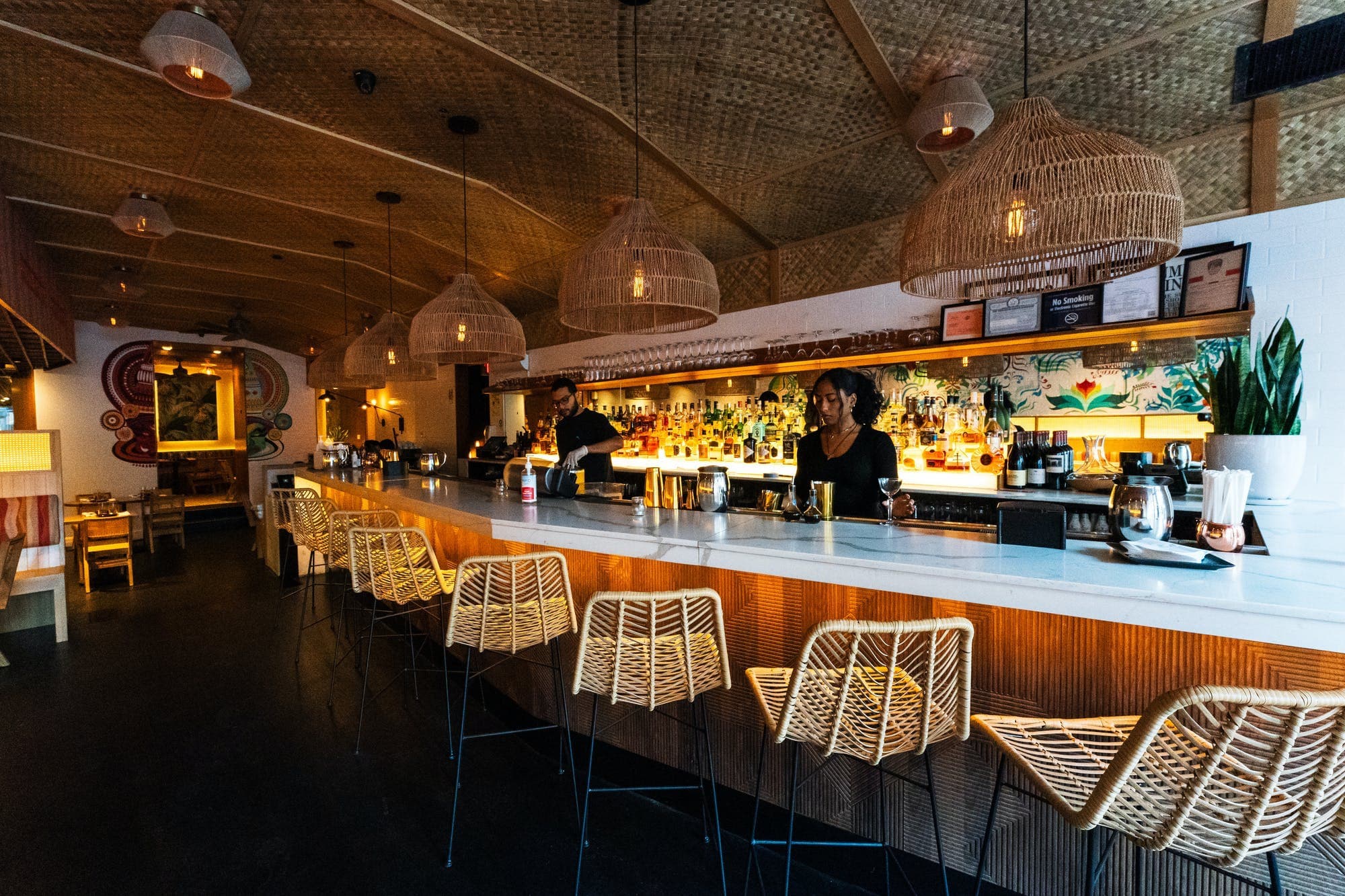 A cozy and inviting bar scene inside Semma restaurant in New York, with bartenders preparing for service. The bar area is illuminated by warm lighting from pendant lamps with large, wicker shades that contribute to the relaxed, bohemian vibe of the space. Woven bar stools line the front of the white bar counter, which is topped with a sleek, dark surface. The backdrop is a lively, tropical-inspired mural that complements the earthy tones and textures of the decor. The ceiling's textured pattern adds depth to the atmosphere, inviting guests to unwind and savor the experience.