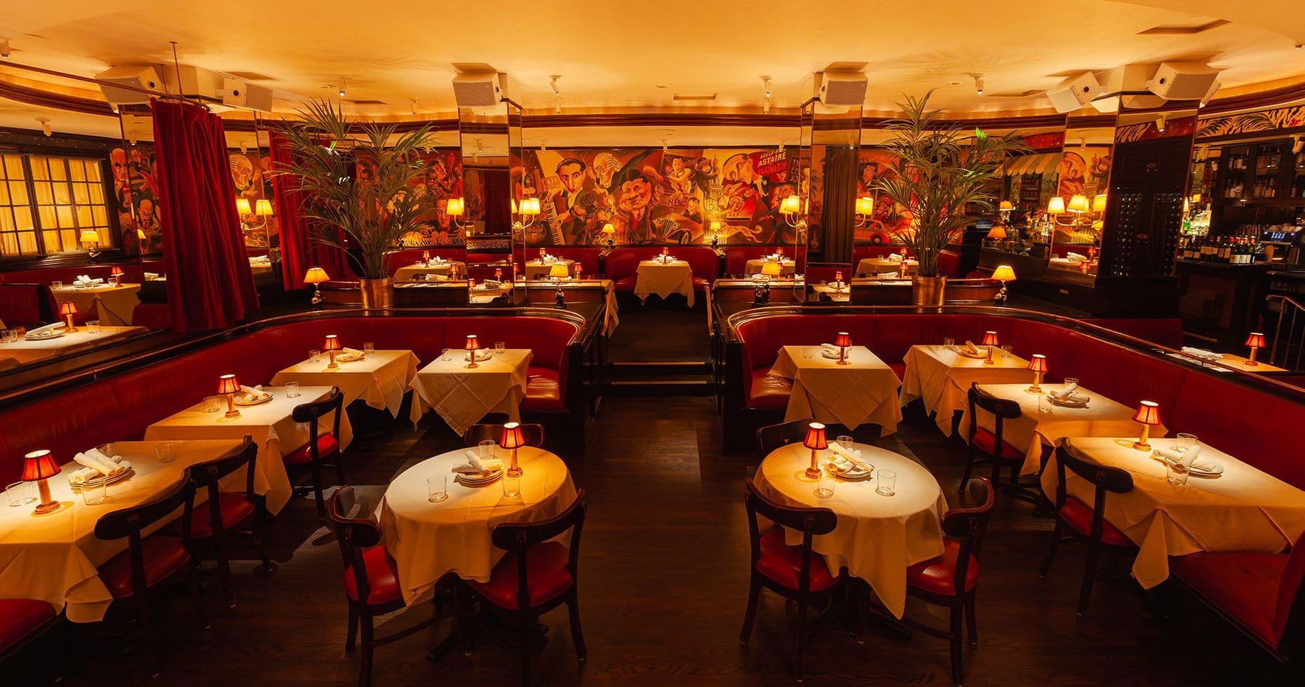 A captivating image of the Monkey Bar restaurant in New York, radiating a warm and intimate ambiance. The room is filled with round tables covered with yellow tablecloths, each set with white plates and napkins, and lit by small, elegant table lamps with red shades. Lush green plants are interspersed throughout, adding a vibrant touch to the rich red leather banquettes that line the walls. The walls themselves are adorned with a large, striking mural depicting an animated, vintage social scene. Soft, ambient lighting complements the dark wooden floors, creating a cozy, retro-glam dining atmosphere.