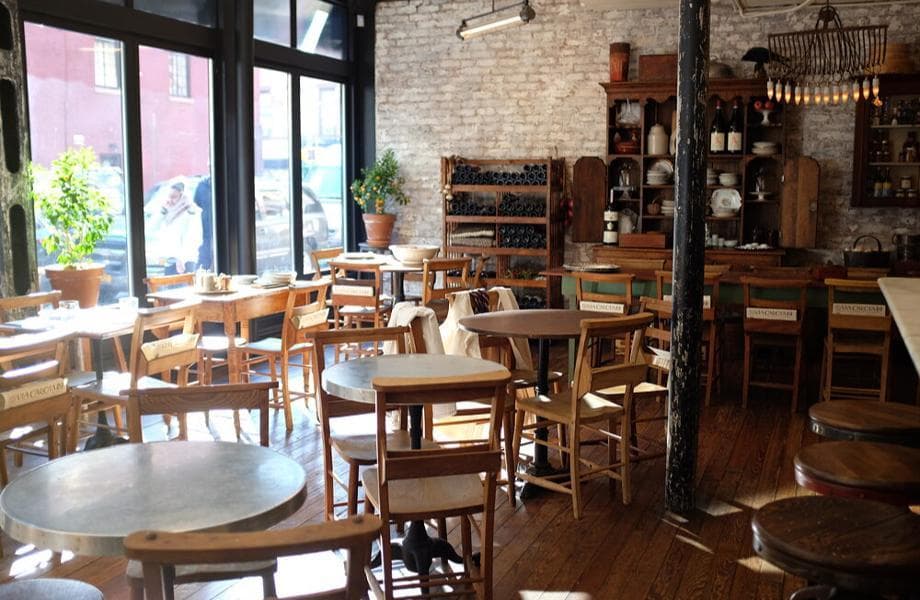 The inviting interior of Via Carota restaurant in New York, bathed in natural light from large windows. The rustic charm is evident in the exposed white brick walls, warm wooden floors, and varied wooden tables and chairs. Shelves along the wall display an assortment of pottery and wine bottles, enhancing the homey atmosphere. In the center, a column, showing signs of its age with peeling paint, adds character to the space. The setting is casual yet stylish, with a few plants providing a splash of greenery, complementing the earthy tones of the decor.