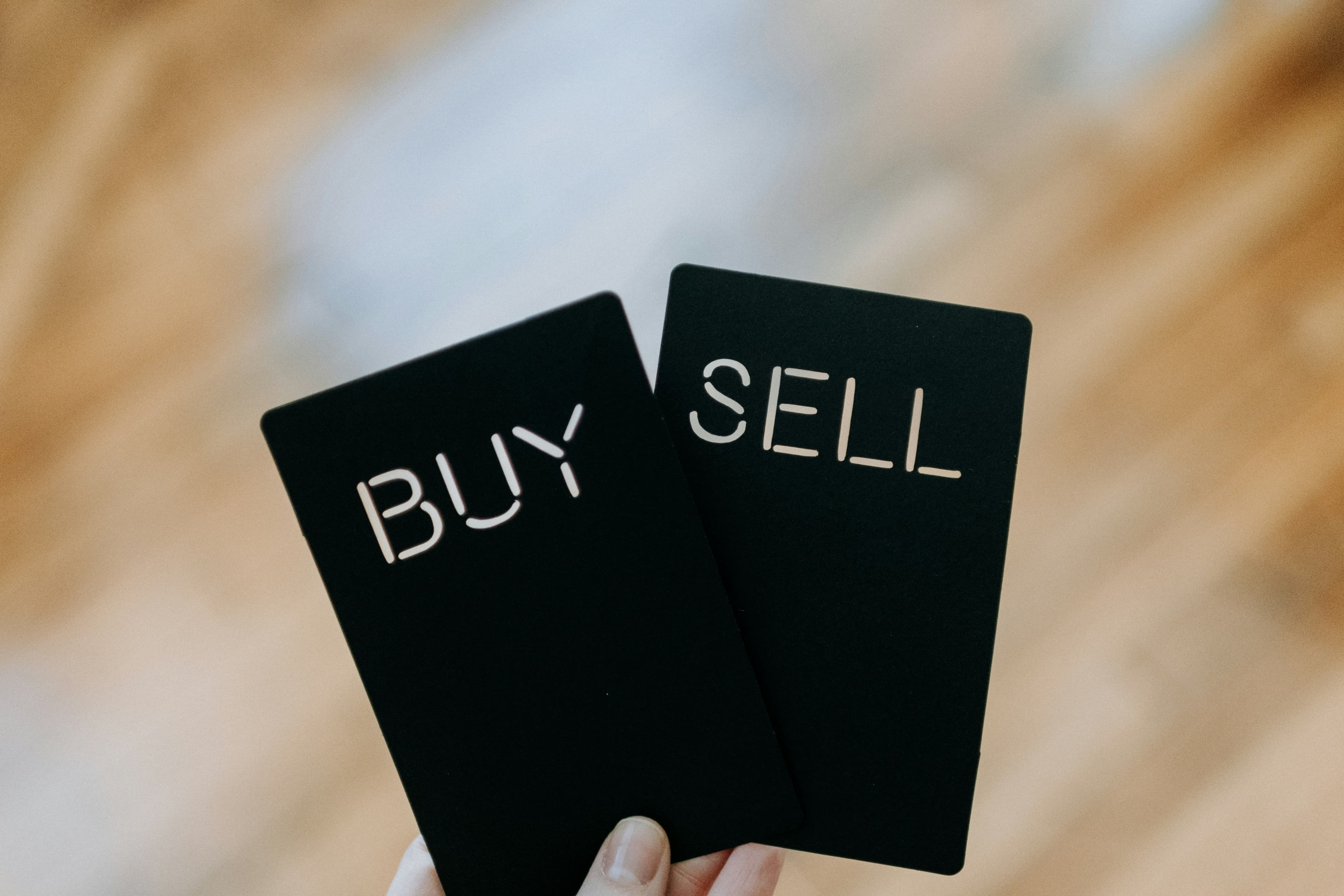 Two hands holding black cards with the words 'BUY' and 'SELL' in bold white lettering, symbolizing the decision-making in trading and commerce against a blurred wooden background.