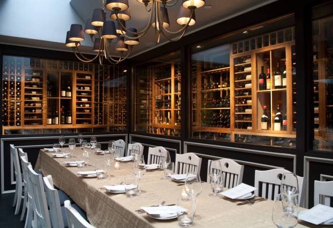 A sophisticated private dining area within L'Artusi restaurant, featuring a long, communal-style table set with crisp white plates and wine glasses. The elegant space is highlighted by a striking wine storage wall, showcasing rows of meticulously arranged bottles in a warmly lit, glass-enclosed cabinet. Above the table, a chic chandelier with multiple round lights casts a soft glow, adding to the ambiance. The room's modern decor, framed by large windows and accented with black trim, creates an intimate atmosphere for a refined dining experience.