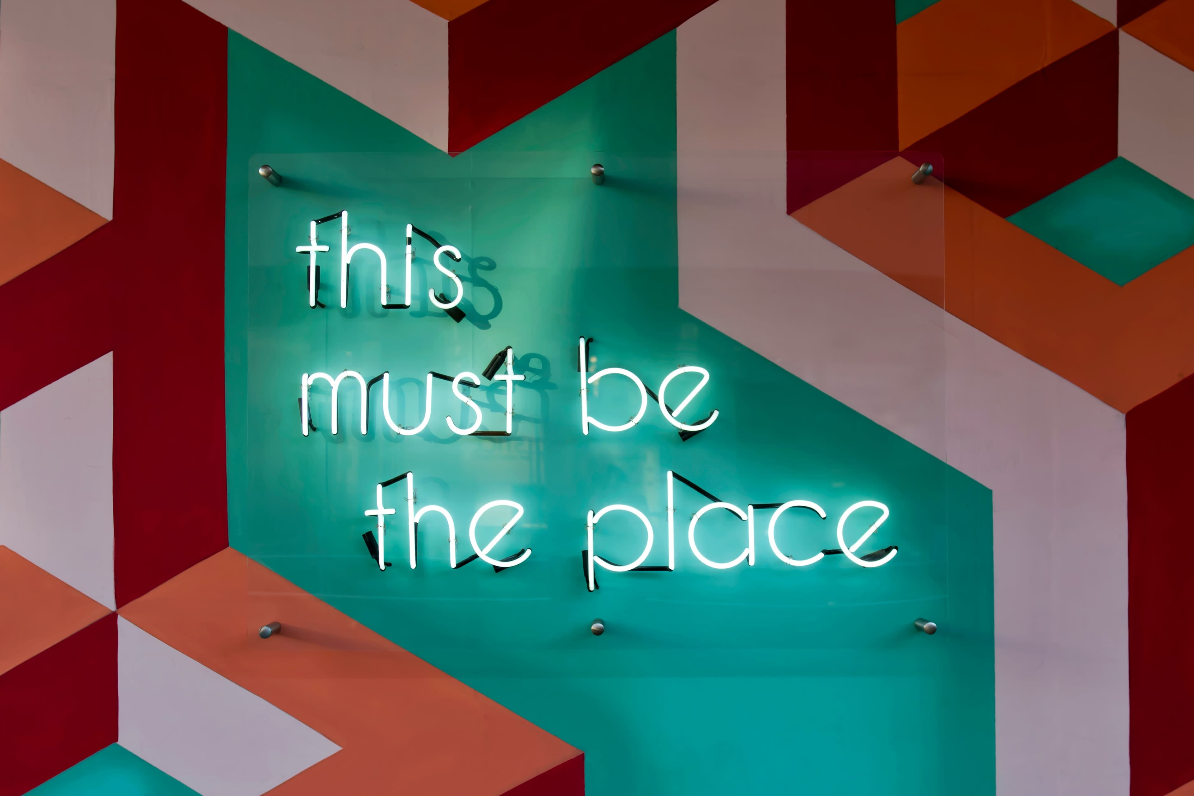 A vibrant and modern art installation, this image shows a neon sign with the cursive text 'this must be the place' glowing in a cool, teal hue against a geometric, multicolored backdrop with a pattern of interlocking red, orange, and white chevrons on a seafoam green wall. The playful and inviting sign serves as both a statement piece and a warm welcome, suggesting a location of interest or a destination point within an urban setting.