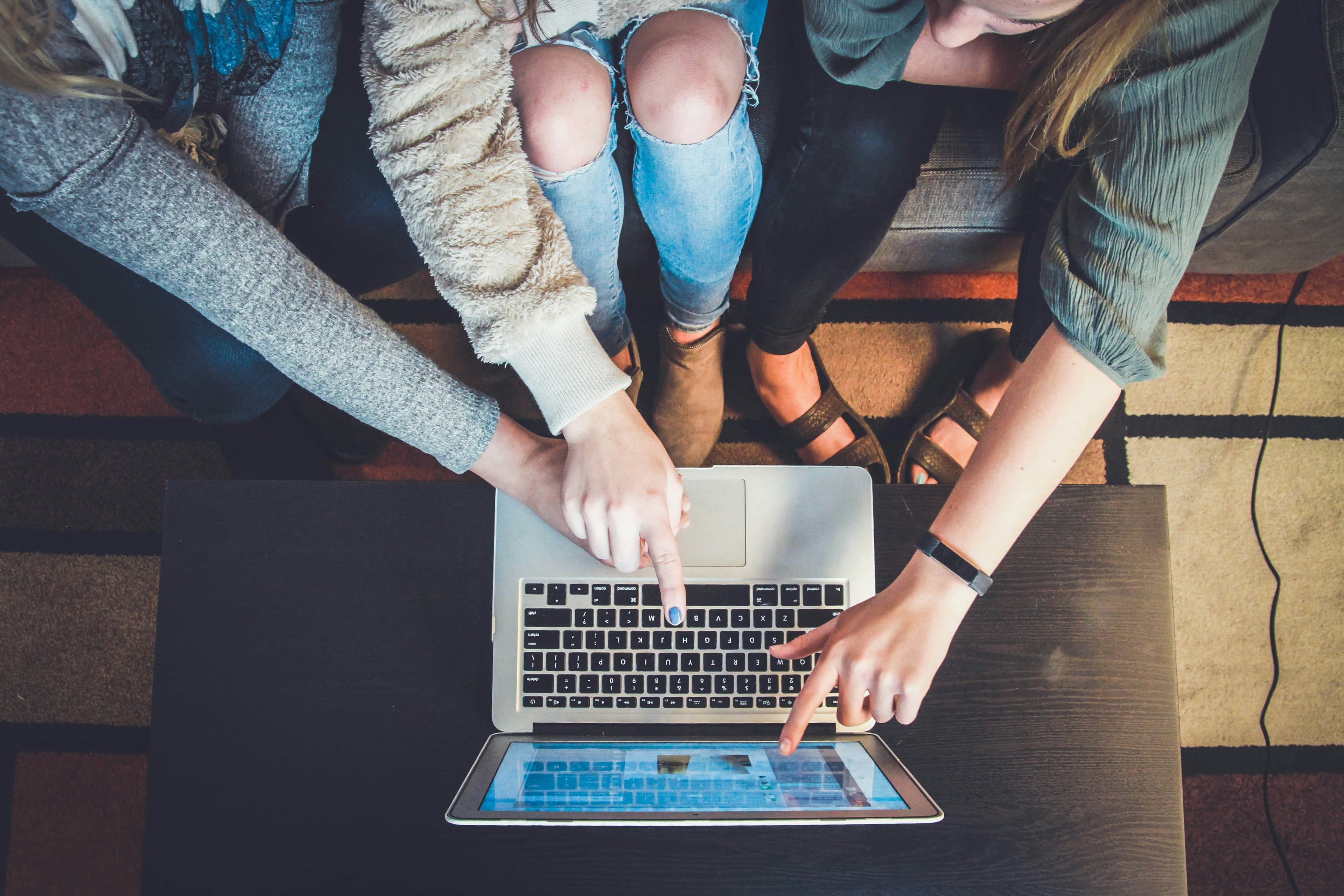 A top-down view of three people gathered around a laptop, with one person pointing at the screen, indicative of a collaborative online search or digital interaction, set against a backdrop of a geometric carpet and casual attire reflecting a relaxed yet engaged atmosphere.