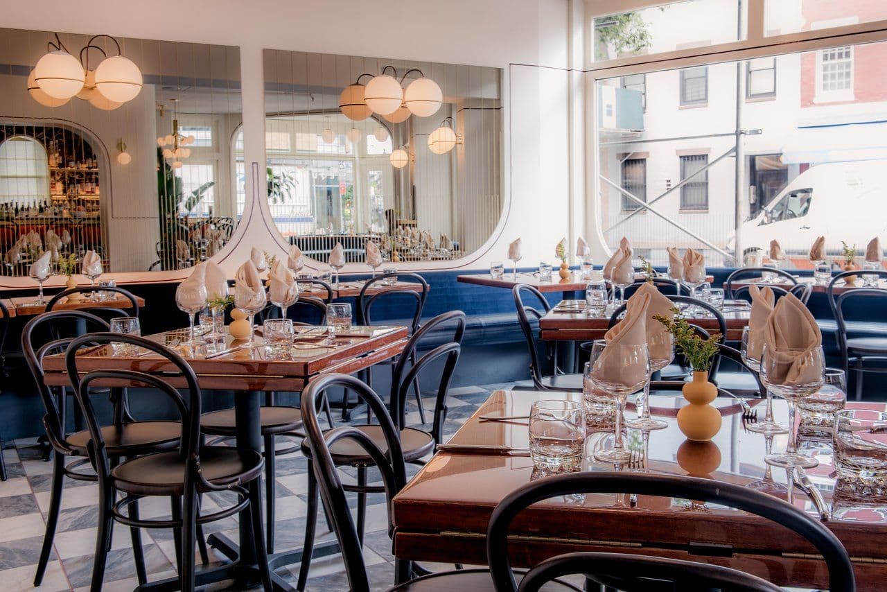 An elegant and brightly lit restaurant interior with a view towards the street. The dining area features neatly arranged tables with glossy wooden surfaces, surrounded by stylish metal chairs with dark, curved backrests. Each table is set with fine glassware and neatly folded beige napkins, creating a sense of anticipation for an upscale dining experience. Overhead, spherical pendant lights cast a warm and inviting glow. The decor includes a long blue upholstered bench against a mirrored wall that reflects the restaurant's charming ambiance. The checkered floor adds a classic touch to the modern and sophisticated space.
