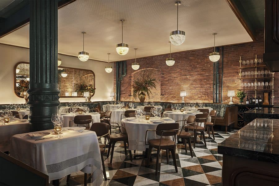 A tastefully decorated restaurant interior combining classic charm with modern elegance. The room is anchored by a striking dark green support column that matches the checkered black and white tiled floor. Round tables draped with crisp white linens are neatly arranged, each set with fine glassware and silverware. The warmth of the exposed brick walls is accentuated by the soft lighting from stylish pendant lamps. Mirrors with arched tops reflect the ambient light and add depth to the space. A bar with a marble countertop and shelves stocked with bottles adds a focal point, while potted plants bring a touch of greenery, enhancing the inviting atmosphere of the dining area.