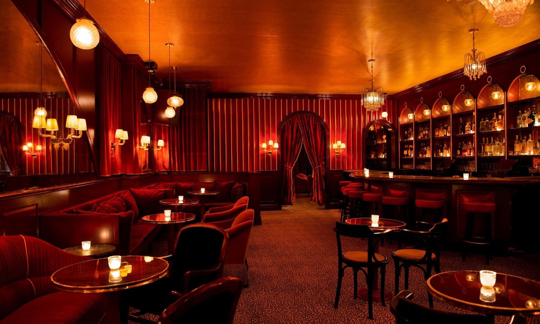 An opulent lounge area that exudes a classic, speakeasy-style luxury with a modern twist. The room is drenched in rich, red hues from the velvet upholstered furniture to the sumptuous drapery. Golden ambient lighting reflects warmly off the gilded ceiling, enhancing the sultry atmosphere. Circular tables with polished tops are scattered throughout, each hosting a flickering candle, adding to the intimate glow. A sophisticated bar area is lined with high stools and backed by a curved, dark wood shelving unit displaying an array of spirits under elegant chandeliers, inviting guests to indulge in a timeless nightlife experience.