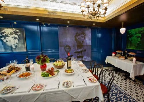 A vibrant dining room exuding an eclectic mix of elegance and artistic flair. The space features deep blue walls with elaborate white trim and a luxurious golden ceiling. A long white tablecloth-covered table is set with colorful patterned plates, candles, and an array of Mediterranean dishes, inviting a festive group dining experience. Prominent black and white portraits add a dramatic touch, with one large image serving as a backdrop that draws the eye. The room is lit by an ornate chandelier, casting a cozy glow over the scene, and the floor boasts a decorative tile design, contributing to the room's dynamic aesthetic.